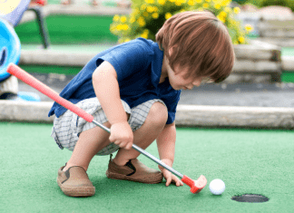 A young boy crouches down on a mini golf lane with his club, pushing the golf ball into the hole.