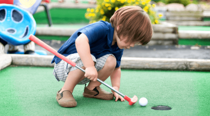A young boy crouches down on a mini golf lane with his club, pushing the golf ball into the hole.