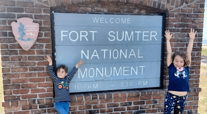 Two children excited with their hands up, standing on either side of the Fort Sumter National Monument sign.