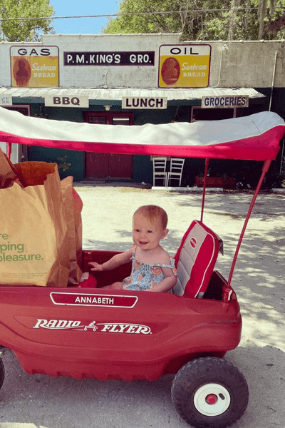 A toddler sitting in a red wagon in front of PM Kings on Wadmalaw Island.