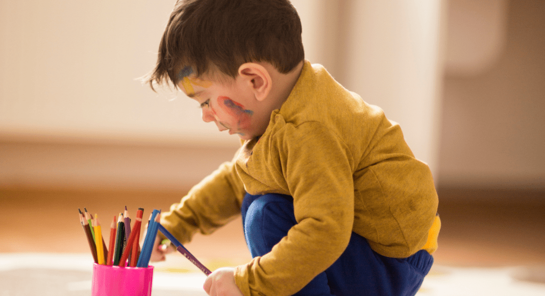 World Poetry Day: a preschool-aged boy crouches down on a giant piece of paper laying on the floor. He has a cup full of colored pencils with one in hand, and splashes of color scribbled on his cheek.