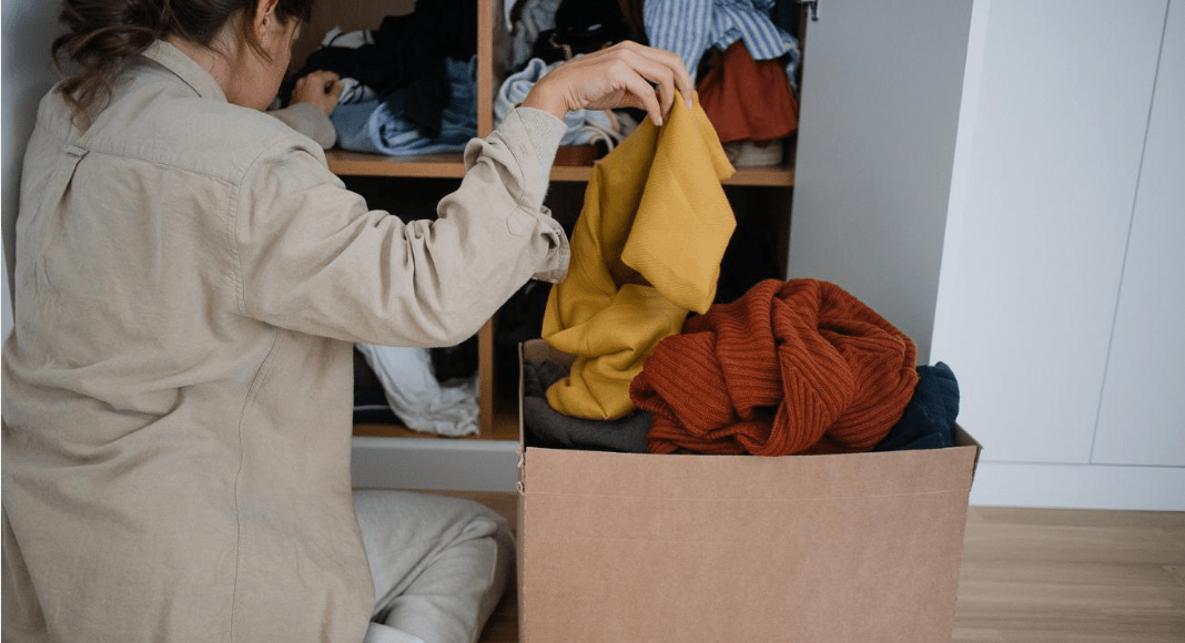selling house: a woman sits on the floor in front of a closet of clothes, dropping a clothing item into a cardboard box.