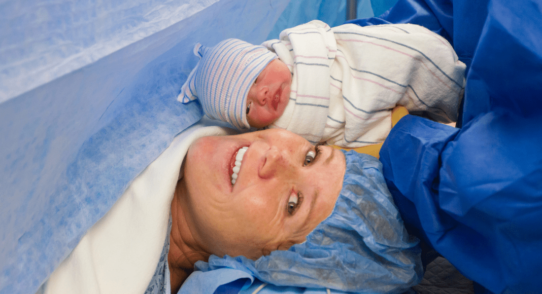 c-section scar: a swaddled baby is held up to her mother's face in front of the blue curtain used during a c-section.