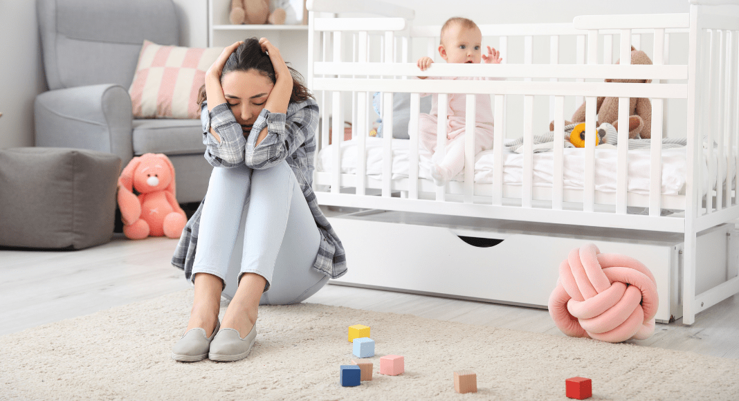Momposter syndrome: A mom sits on the nursery floor with her head on her knees, while her baby is sitting up in the crib behind her.