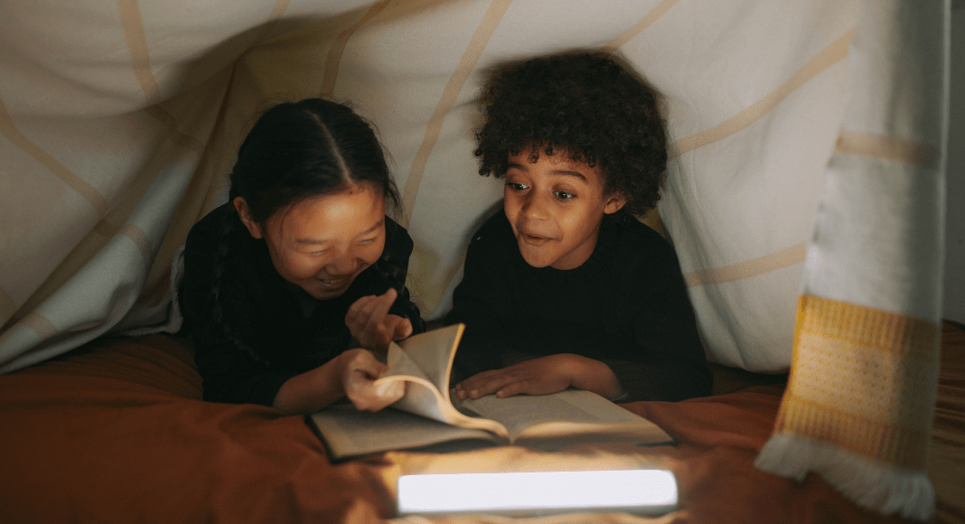 a school-aged girl and boy are under a blanket fort flipping through a book together.