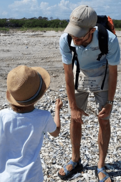 guided shark tooth hunting: a man tour guide leans over listening to a small child. They're standing on a rocky beach with blue skies overhead.