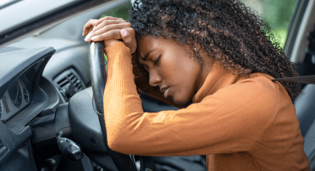 Nowhere feels safe: a woman sits in a car holding the steering wheel, with her head against her hands. Her eyes are closed and she has a concerned expression.