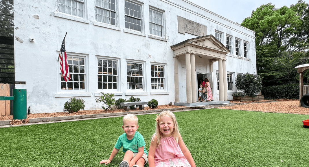 Two children sitting on the grass in front of a school building.