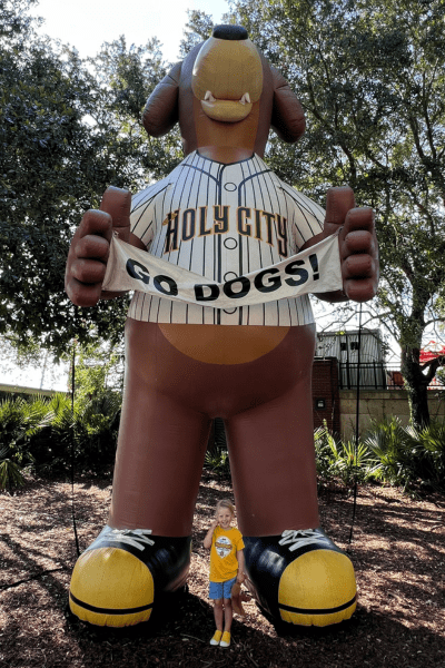 A giant brown inflatable dog wearing a white Holy City baseball jersey, holding a sign that exclaims, "Go Dogs!" A young girl in yellow and blue stands in front.
