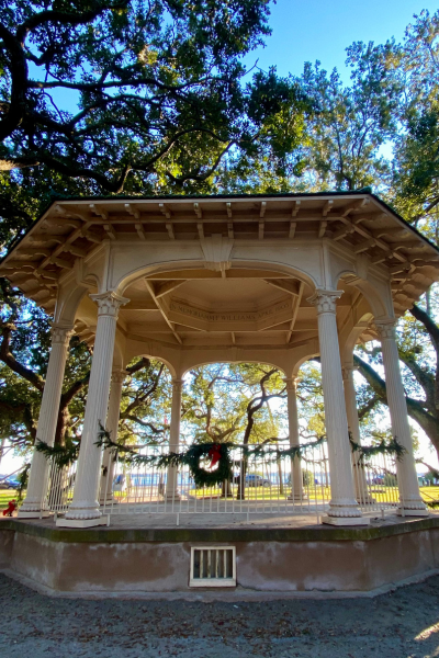 A white gazebo with greenery around its railings sits under large trees and a bright blue sky.