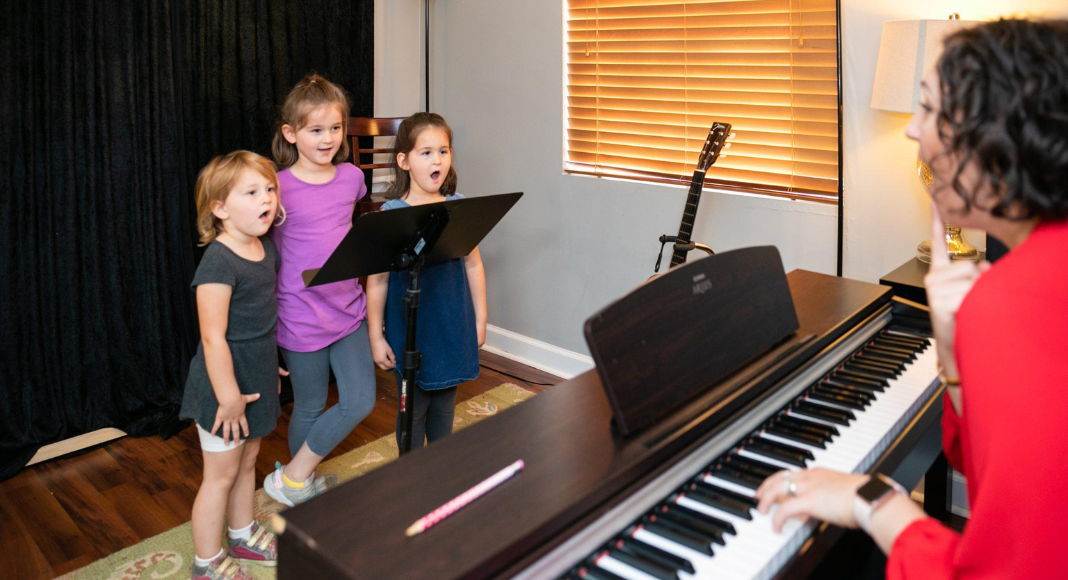 A music lesson teacher sits at a piano and instructs three young girls while they sing.