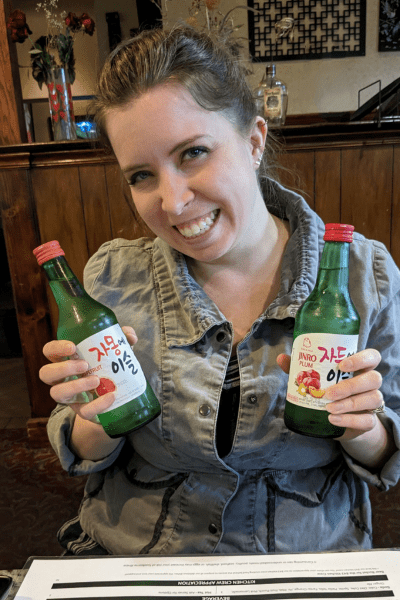 A woman smiles, holding two different soju drink bottles.