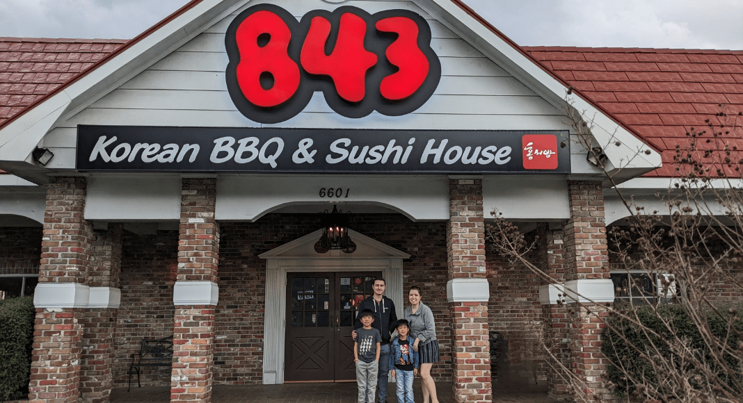Outside view of 843 Korean BBQ & Sushi House with a family of four standing beneath the sign.