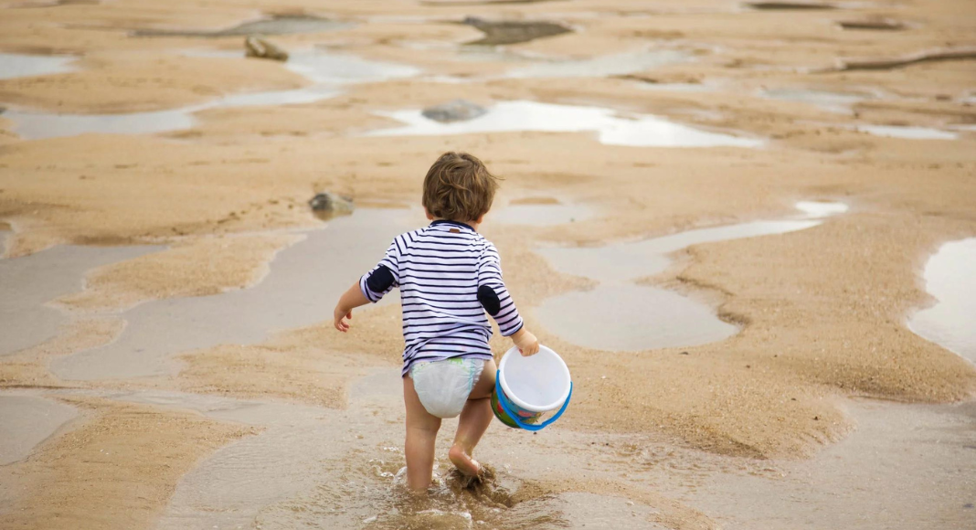 beach outing with kids: a toddler walks in shallow tide pools with a bucket in hand.