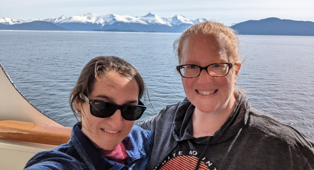 Two women on a ladies' trip, posing on cruise ship with water and white mountains in the background.