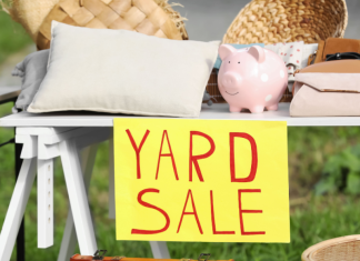 A yard sale sign is hung on the front of a table with lots of stuff on it.