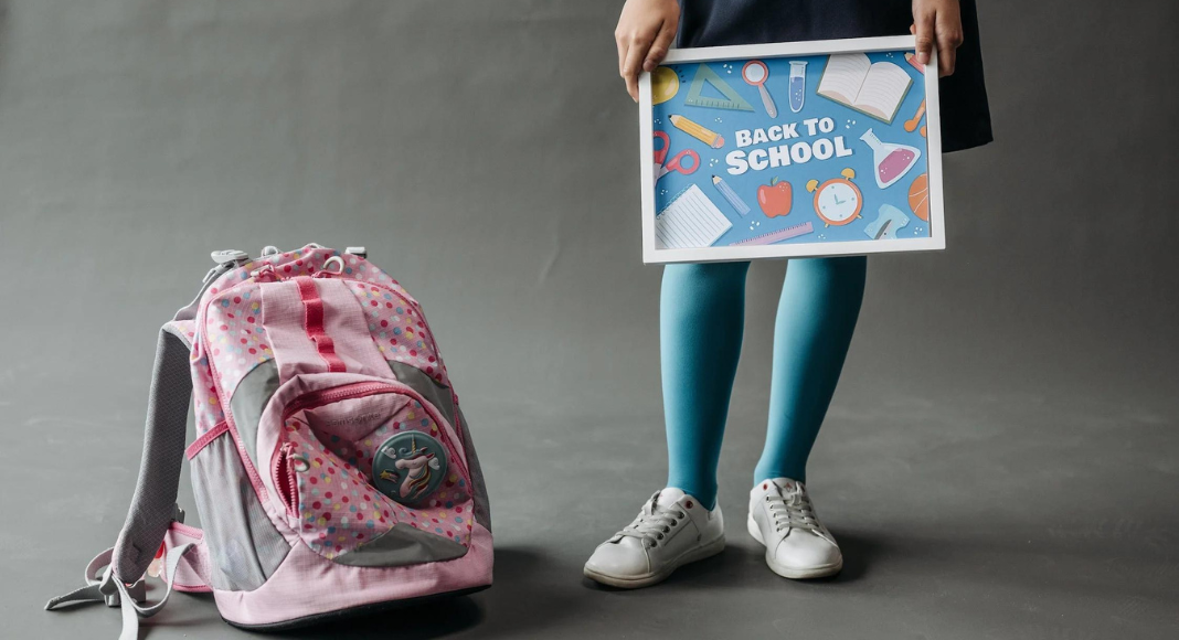 end of summer traditions: a child holds a sign saying "back to school" with her backpack on the floor next to her.