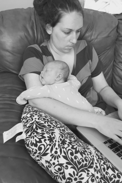 A young mom tries to balance holding her infant on the couch while working on her laptop.