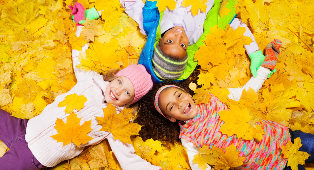 Thanksgiving traditions: 3 children lay in a pile of yellow leaves with their heads together, smiling.