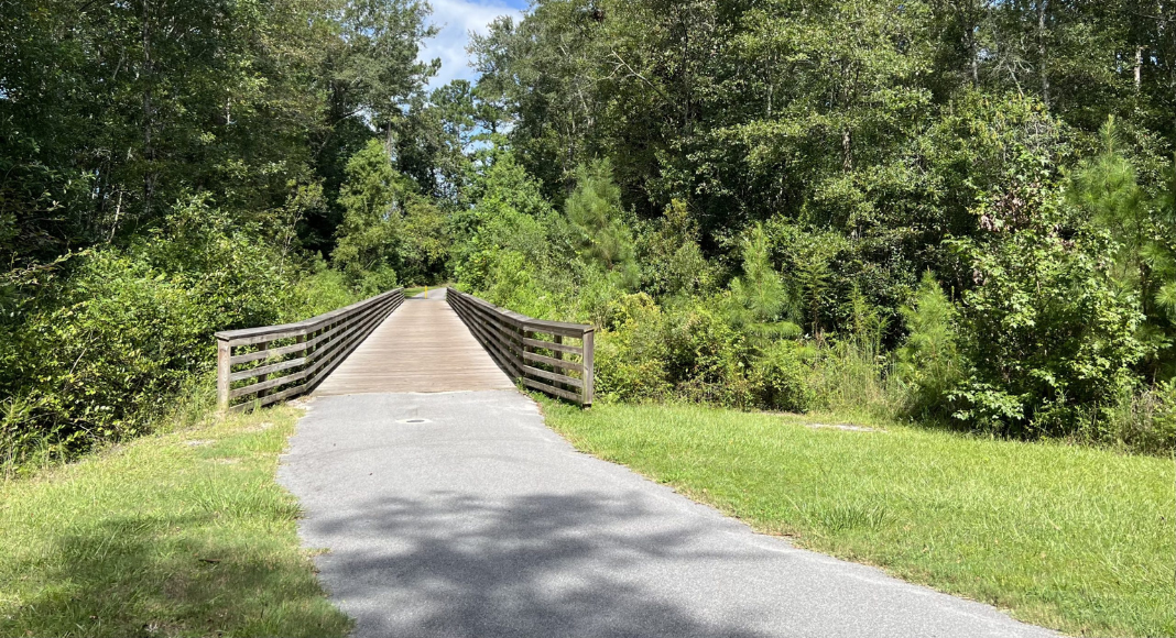 A paved trail leading to a small wooden bridge surrounded by trees and foliage