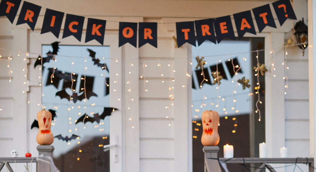 front porch covered with halloween decor: pumpkins, trick or treat banners, paper bats, and string lights.
