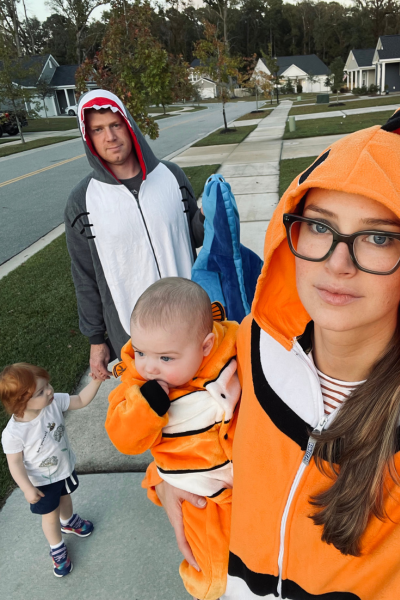 A toddler girl in regular clothes walking with her family all dressed in costumes, with her Halloween costume in her dad's hand.