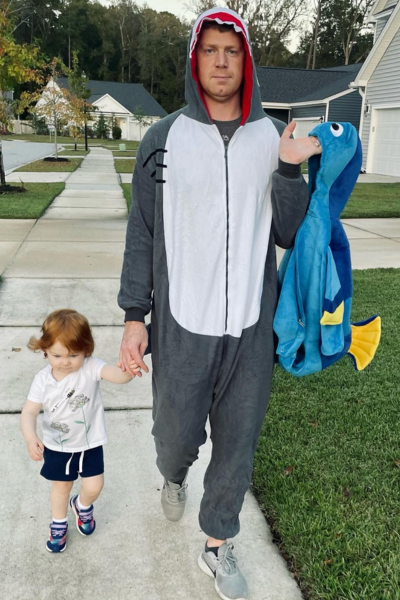 A toddler girl in regular clothes walking with her dad dressed in a shark costume, with her Halloween costume in his other hand.