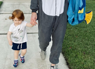A toddler girl holding her dad's hand with her Halloween costume in his other hand.