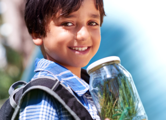 A boy wearing a backpack holds a jar of grass with a plastic grasshopper in it.
