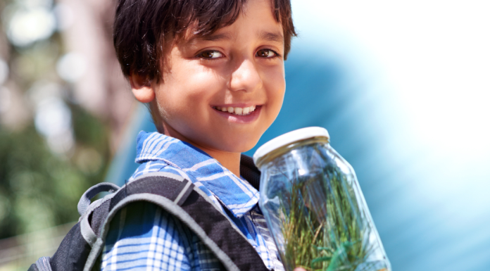 A boy wearing a backpack holds a jar of grass with a plastic grasshopper in it.