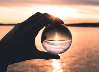 A hand holds a glass orb up to a sunset overlooking a body of water