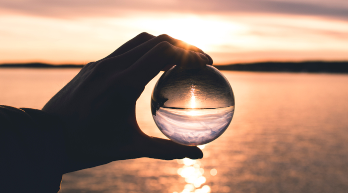 A hand holds a glass orb up to a sunset overlooking a body of water