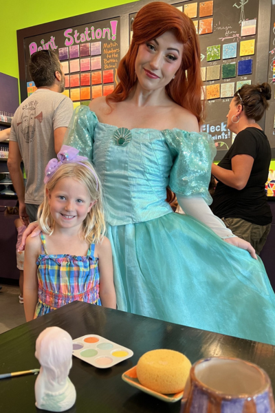 experience gifts for kids: a little girl smiles with Princess Ariel with a pottery painting project on the table in front of them.