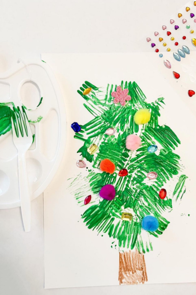 A plastic fork with green paint on the back of its prongs, next to a Christmas tree painting with pom-poms, and jewel stickers decorating it