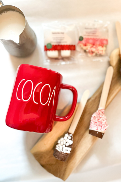 A red mug that says COCOA, with a small pitcher of milk, wooden spoons layered with chocolate and toppings.