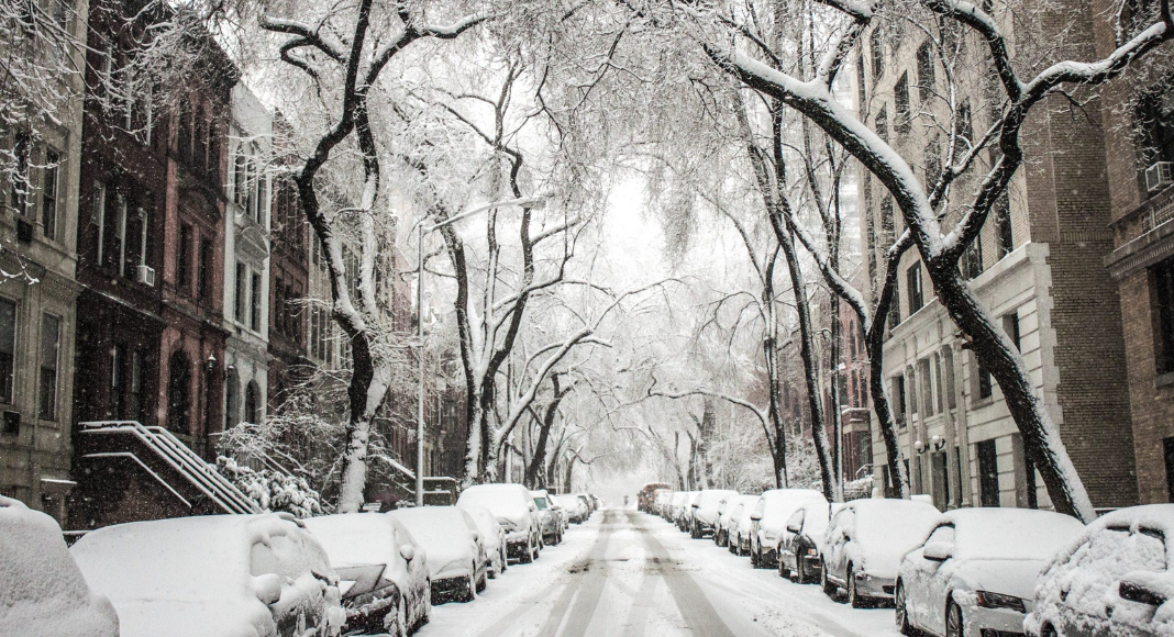 A snowy scene of a urban road lined with cars, trees, and apartments.