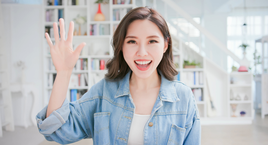 Friendship dating: A woman waves "hello" in front of a large bookshelf.