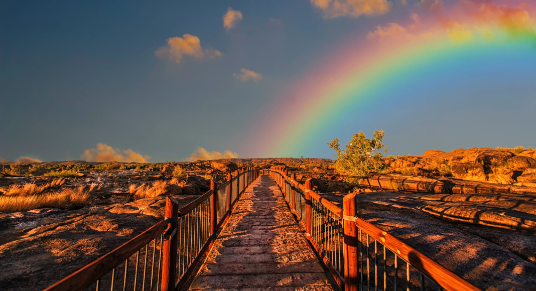 A wooden bridge leads to a sky with a rainbow.
