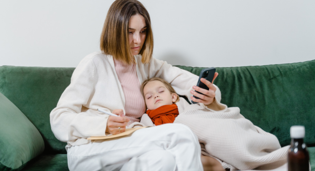 A mother sits on the couch snuggling with her child sick with the stomach bug. The mother looks at her phone and is taking notes.