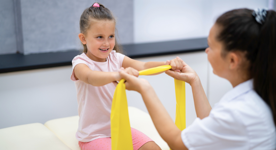 A young girl stretches a band along with her occupational therapist.