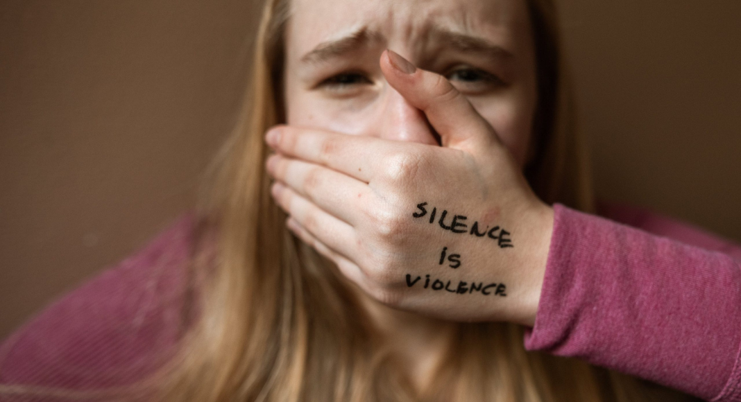 Abuse: a teenage girl covers her mouth with her hand that says "Silence is Violence." She has a hurt, distraught expression.