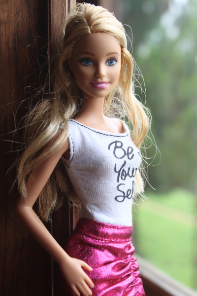 A Barbie doll stands in a window and wears a shirt that says, "Be Yourself."