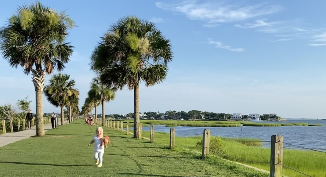 Little girl running across the grass with her teddy bear at Pitt Street Bridge. Palm trees, blue water, and blue skies surround her.