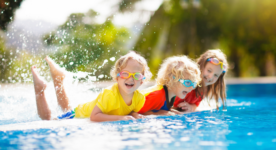 Three kids wearing goggles, splashing water in a pool with their feet.