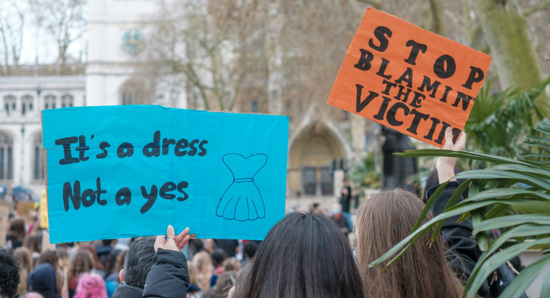 People in a crowd hold up signs that say, "It's a dress, not a Yes." and "Stop blaming the victim."