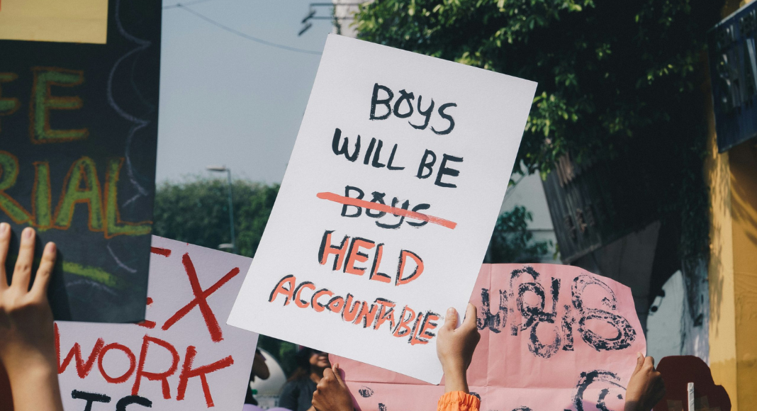 Sexual Assault: Someone in a crowd holds up a sign that reads, "Boys will be held accountable." Instead of, "Boys will be Boys."