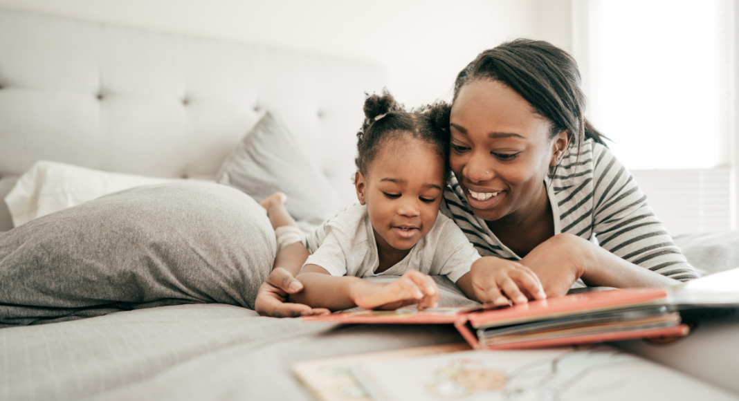 A mom and her young daughter reading a book together in the mom's bed as part of their bedtime routine.