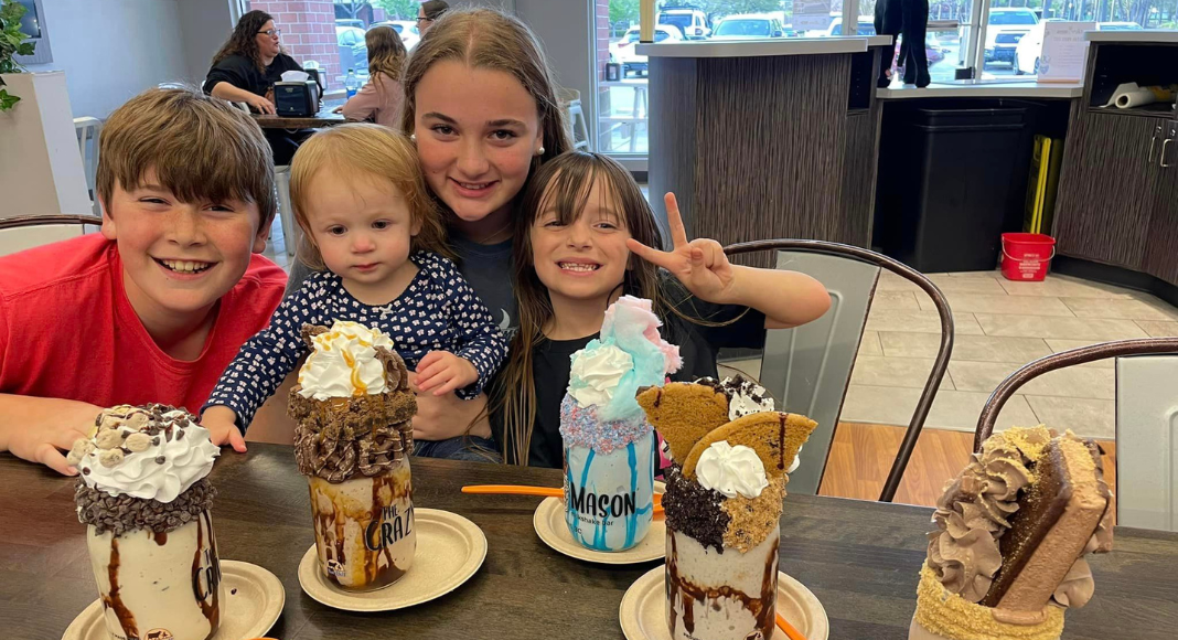 A group of four kids smile together behind four massive milkshake desserts.