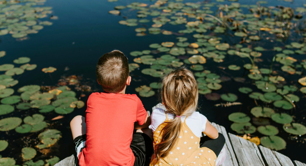 STEM: Two children sit on a dock overlooking a pond with lily pads.