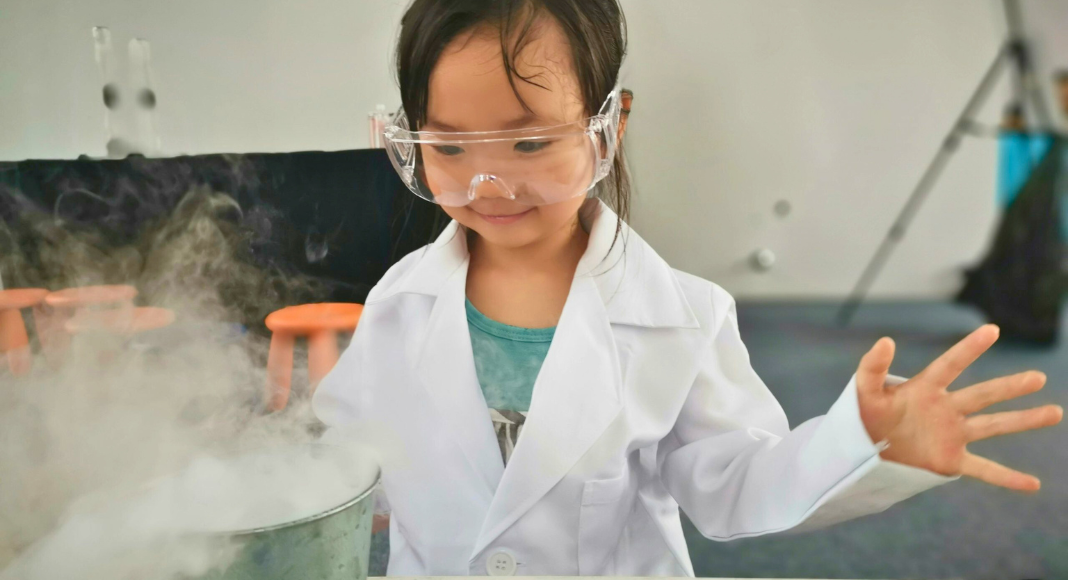 A young girl with safety glasses on peers at a pot with steam coming out of it.